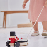 Close up of young girl in a pink dress pulling a PlanToys musical pull along drummer toy on a white floor