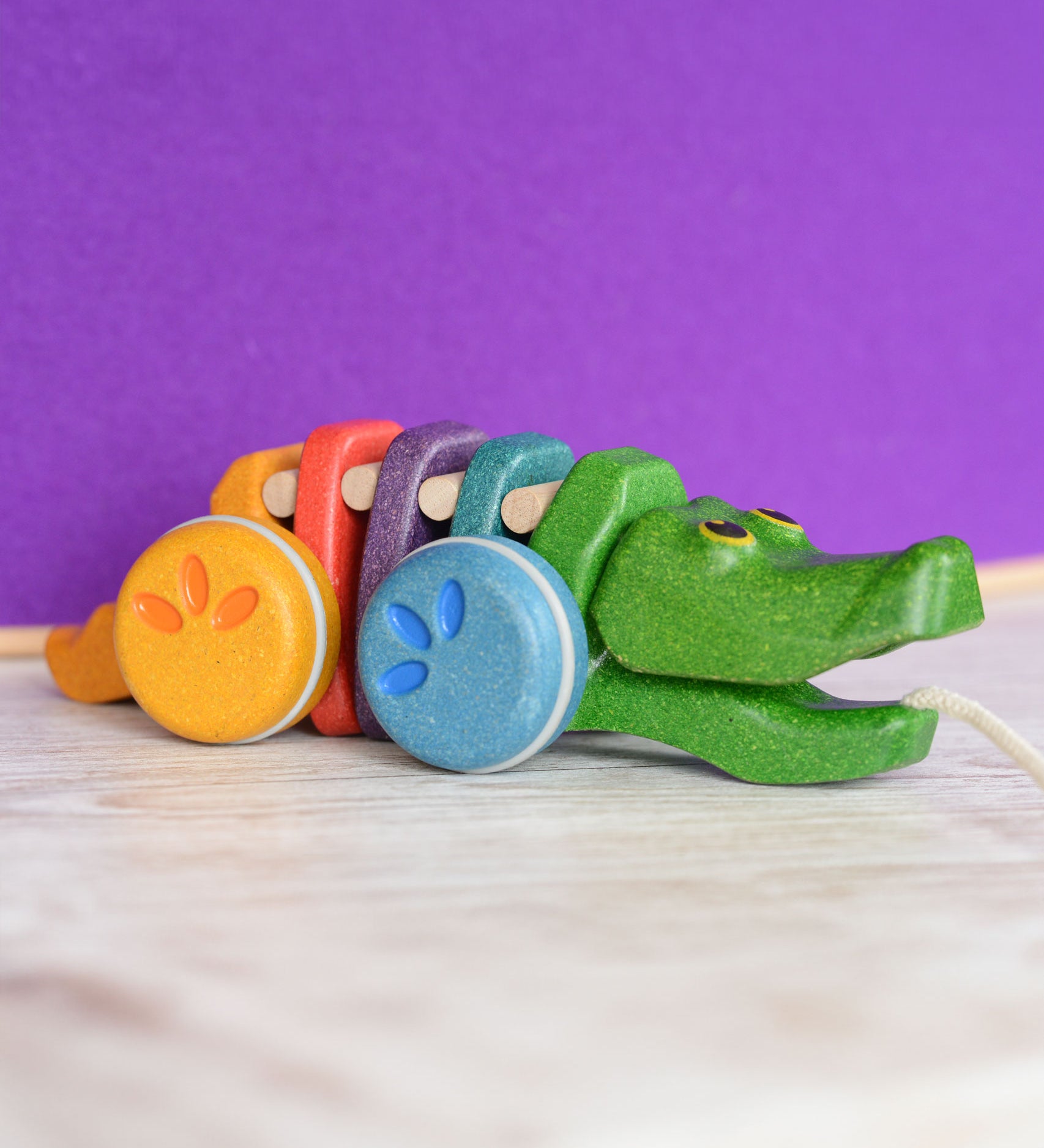 A Plan Toys Rainbow Alligator placed on a wooden surface in front of a purple wall. 