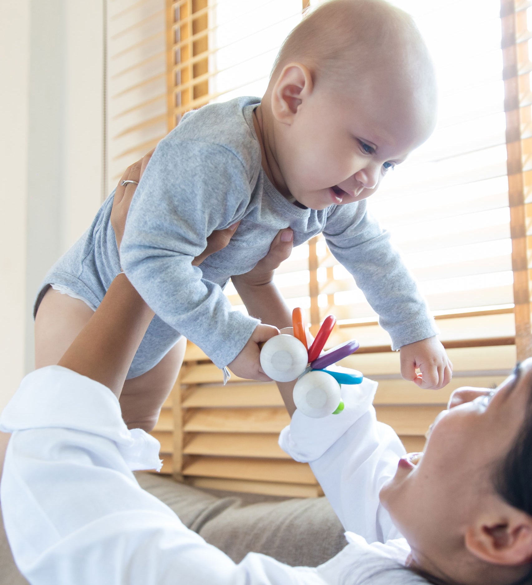 An adult holding up a baby, the baby has a PlanToys Baby Car Rattle in their hand. 