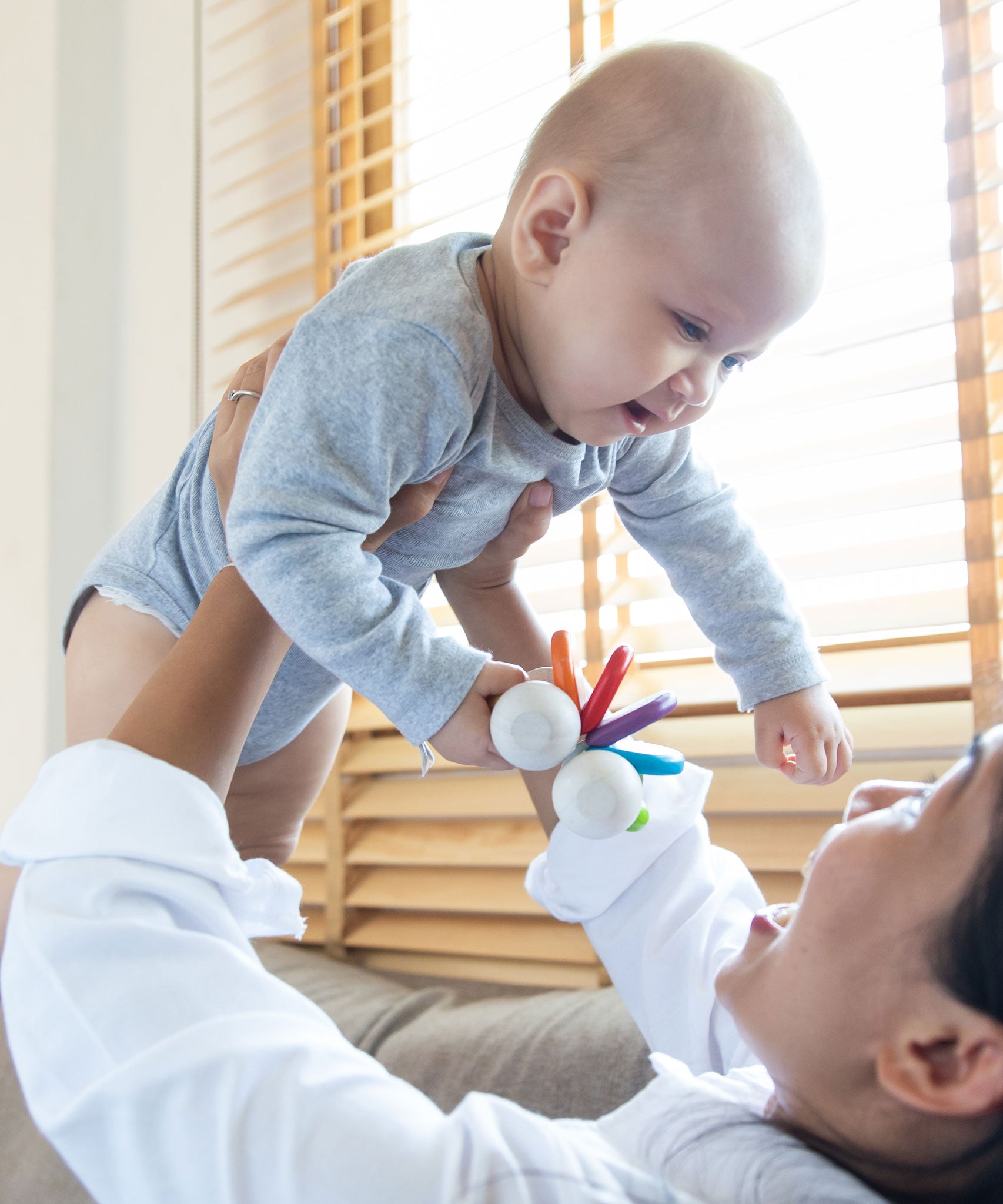 An adult holding up a baby, the baby has a PlanToys Baby Car Rattle in their hand. 