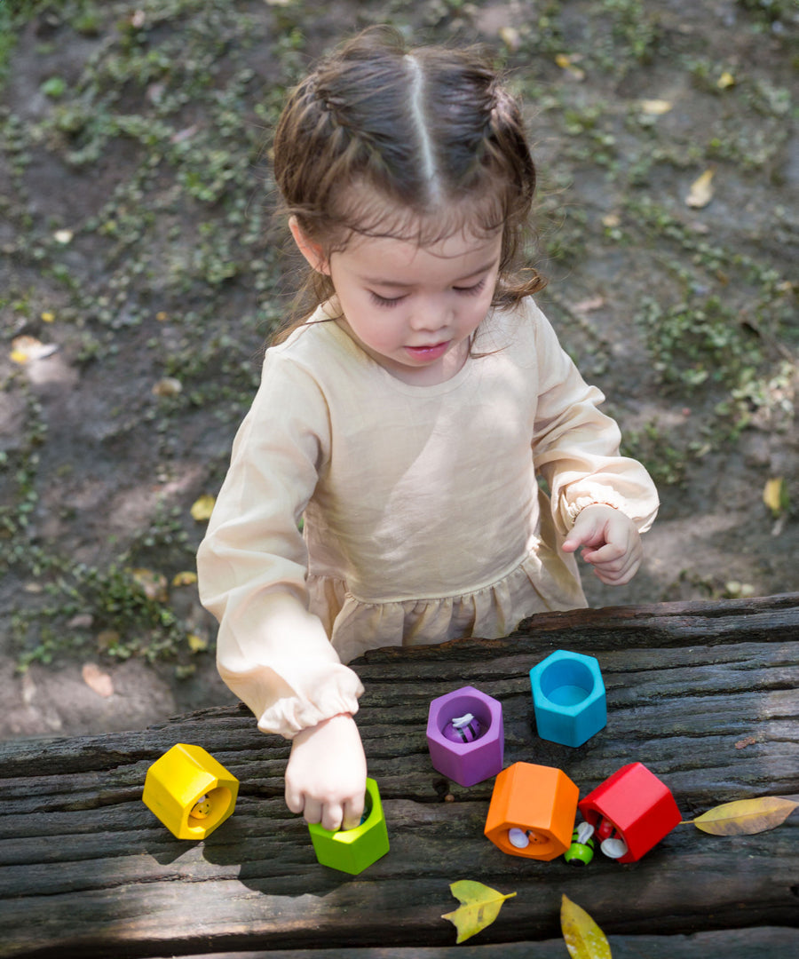 A child playing with the PlanToys Bee Hives Rainbow set outdoors. The bees and hives have been placed on a wooden surface. 