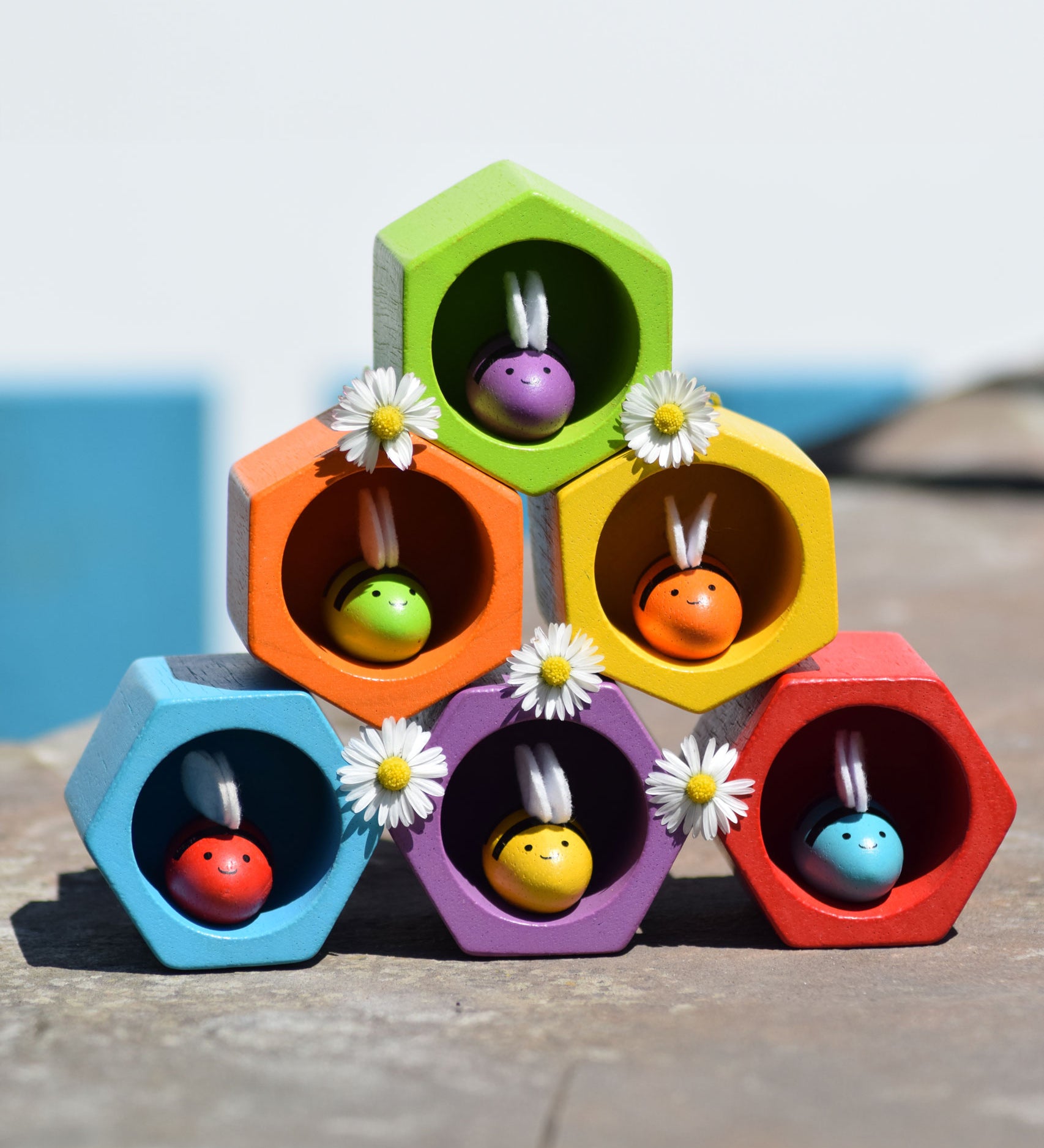 The PlanToys rainbow bee and bee hives set stacked up in a 3, 2, 1 formation. All the bees are in the hives daisies have been placed in-between the hives. 