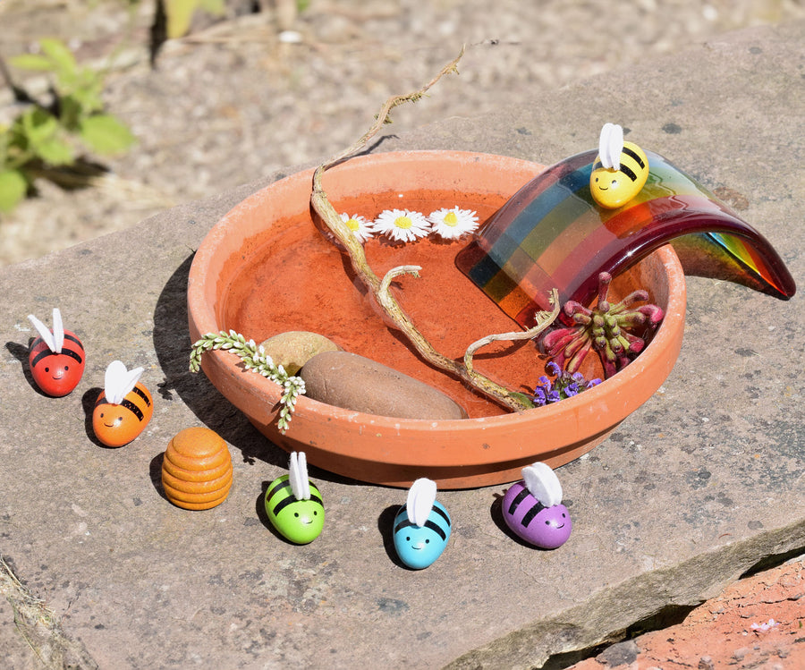 The bees from the PlanToys rainbow bees set being used in a small world play set up outdoors. A terracotta plant stand has been filled with water, some flowers, a stick and a rock. A glass rainbow has been placed on the side with the yellow bee sitting proudly on to.  