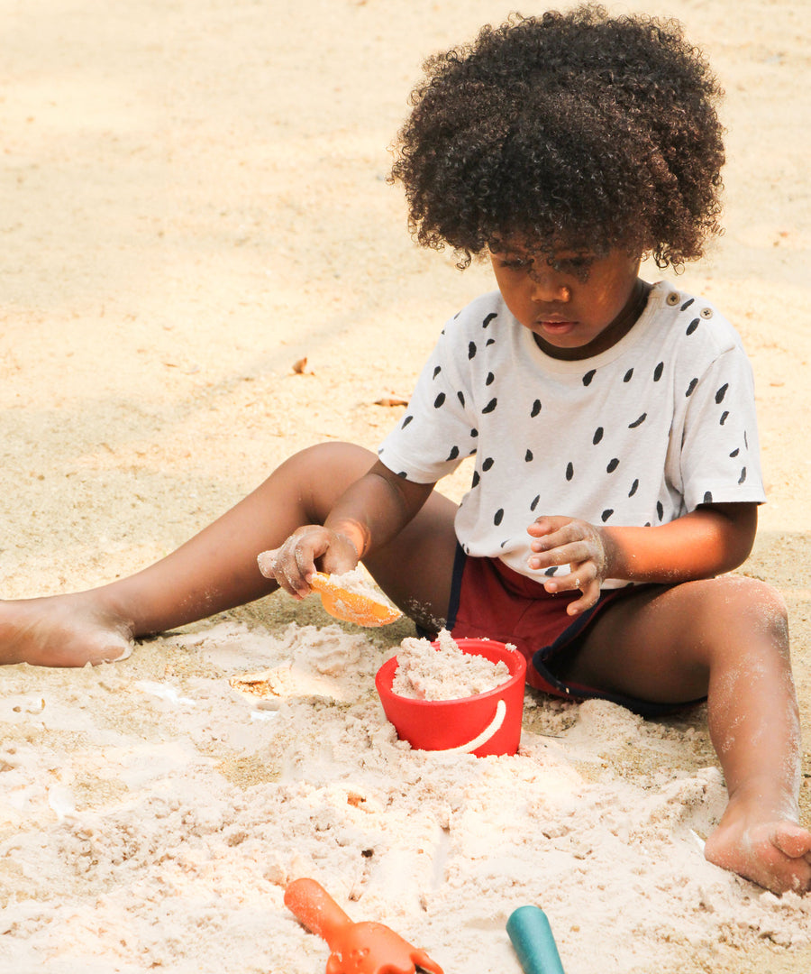 A child filling up the red bucket from the PlanToys Sand Play Set using the yellow tool. 