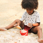 A child filling up the red bucket from the PlanToys Sand Play Set using the yellow tool. 