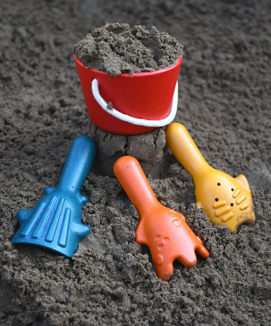 The PlanToys Sand Play Set pictured on dark coloured sand. The red bucket is filled with sand with the three tools placed on the sand in front of the bucket. 