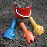 The PlanToys Sand Play Set pictured on dark coloured sand. The red bucket is filled with sand with the three tools placed on the sand in front of the bucket. 