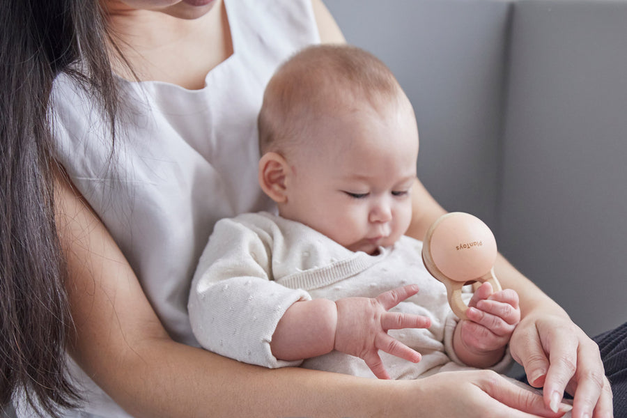 Close up of a baby in a woman's arms playing with the PlanToys plastic-free wooden shake rattle