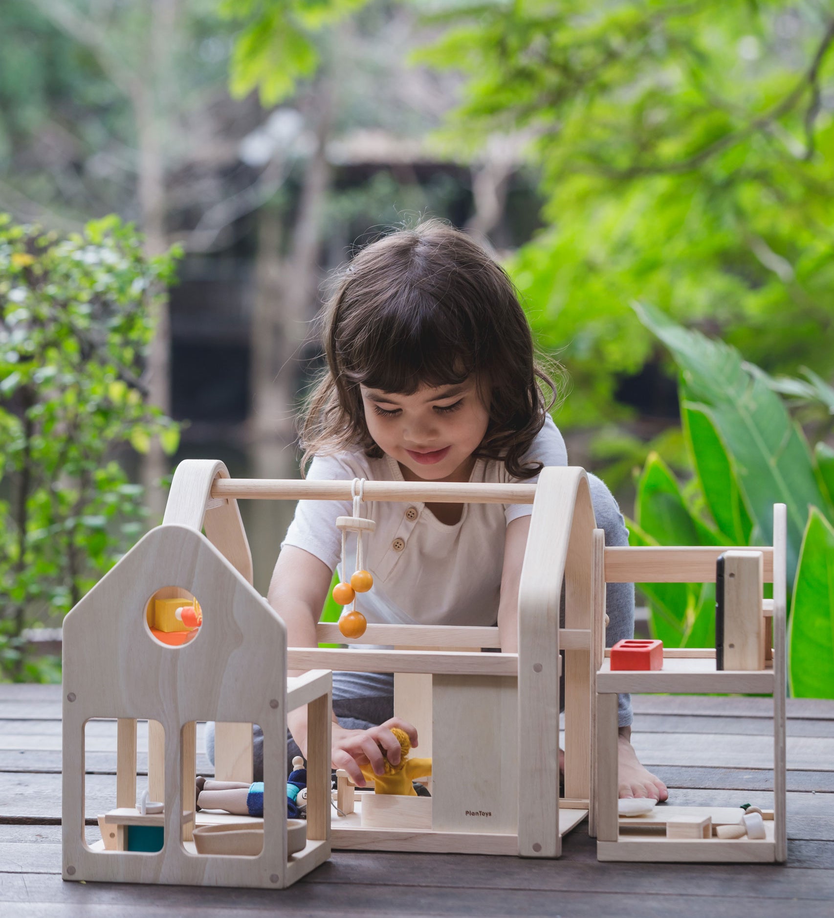 A child sitting on wooden decking outdoors playing with the PlanToys Slide N Go Dolls' House. 
