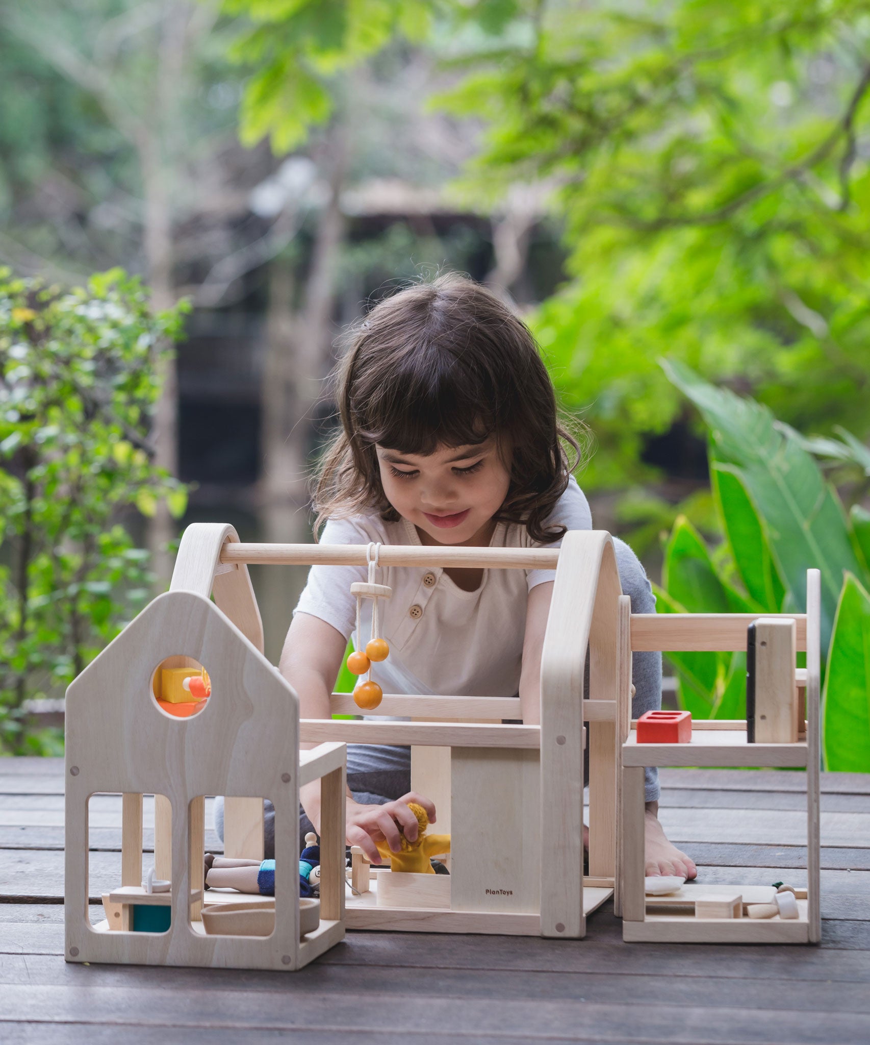 A child sitting on wooden decking outdoors playing with the PlanToys Slide N Go Dolls' House. 
