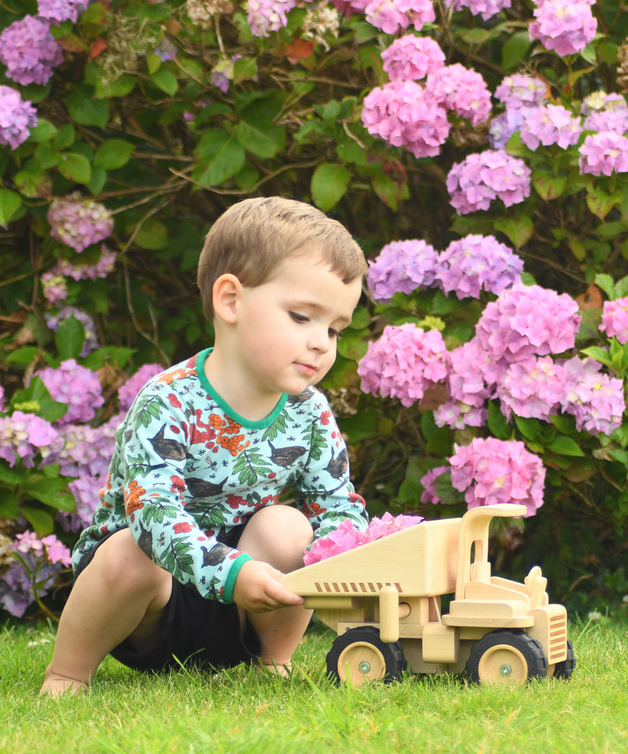 A child playing with the  Plan Toys Special Edition Natural Dump Truck outdoors on grass. There are purple hydrangeas behind them 