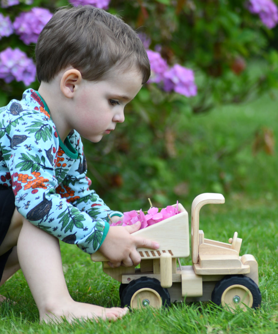 A child playing with the  Plan Toys Special Edition Natural Dump Truck outdoors on grass. There are purple hydrangeas behind them. The child is pushing the dumper truck along with hydrangea petals in the back of the truck. 