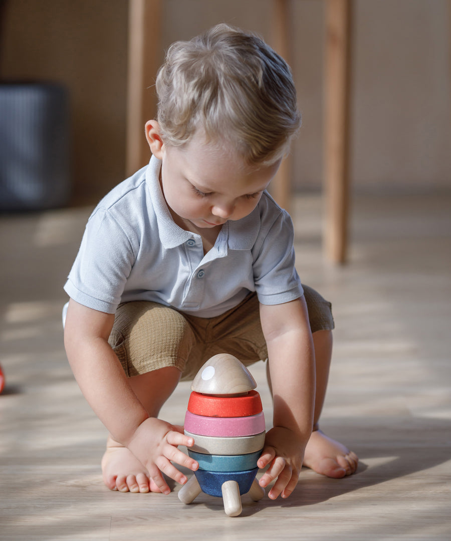 A child crouching down playing with the PlanToys orchard colour way Stacking Rocket Baby Toy.