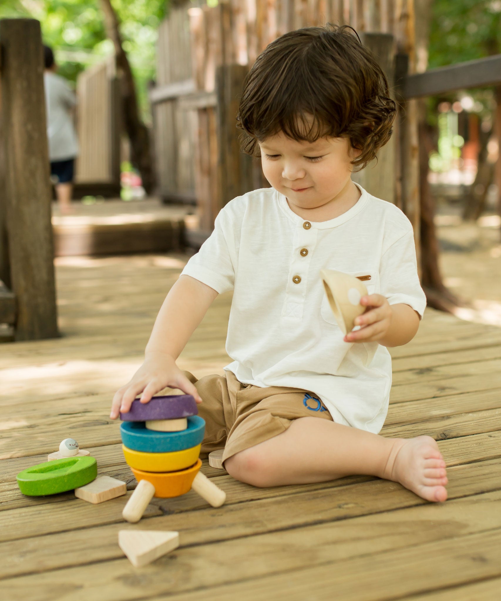 A child sitting on wooden decking outdoors playing with the PlanToys Stacking Rocket toy. 
