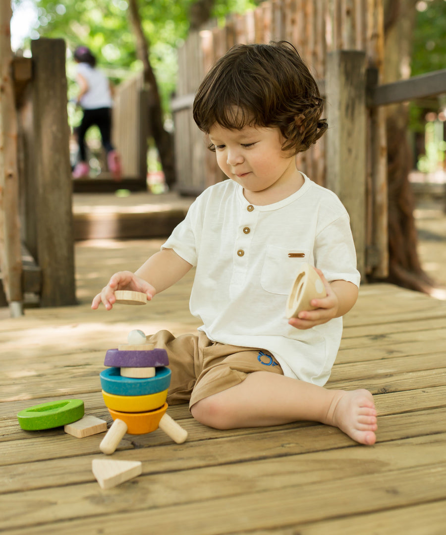 A child sitting on wooden decking outdoors playing with the PlanToys Stacking Rocket toy. 
