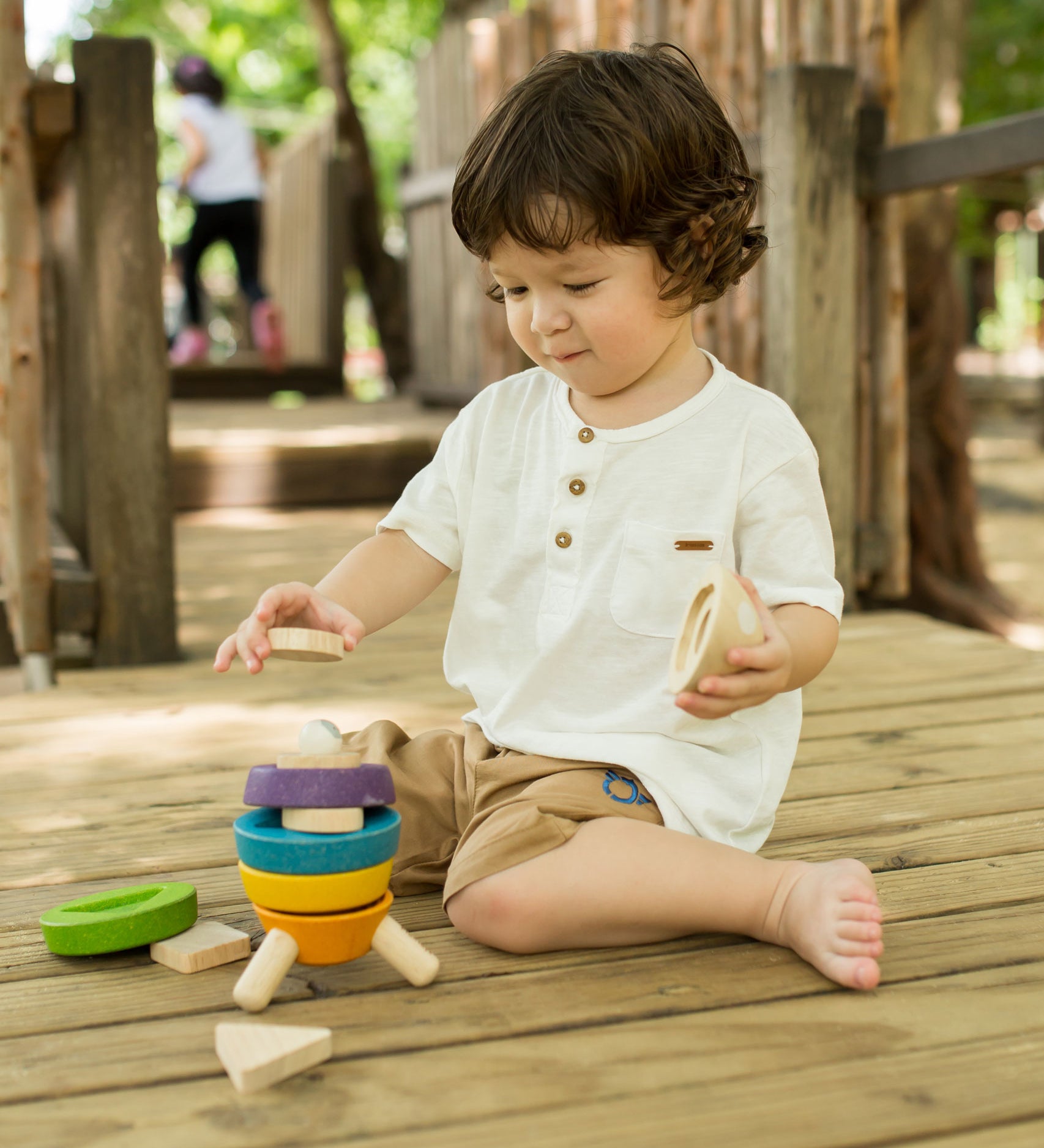 A child sitting on wooden decking outdoors playing with the PlanToys Stacking Rocket toy. 
