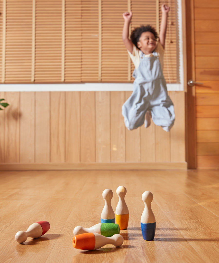 A child playing with the PlanToys Bowling Set indoors on a wooden floor. The child can be seen jumping with joy in the background. 
