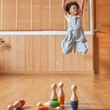 A child playing with the PlanToys Bowling Set indoors on a wooden floor. The child can be seen jumping with joy in the background. 
