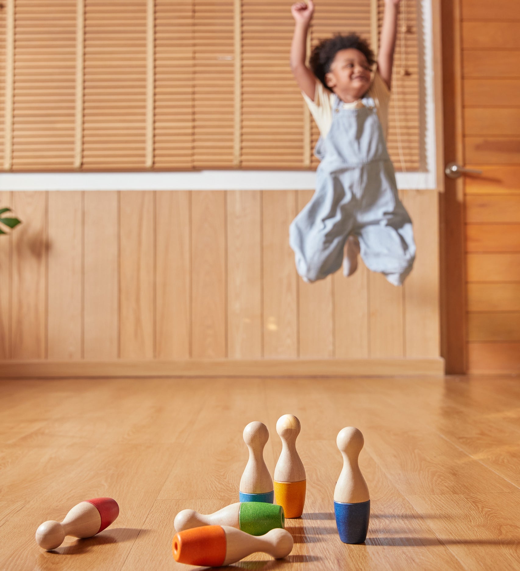 A child playing with the PlanToys Bowling Set indoors on a wooden floor. The child can be seen jumping with joy in the background. 
