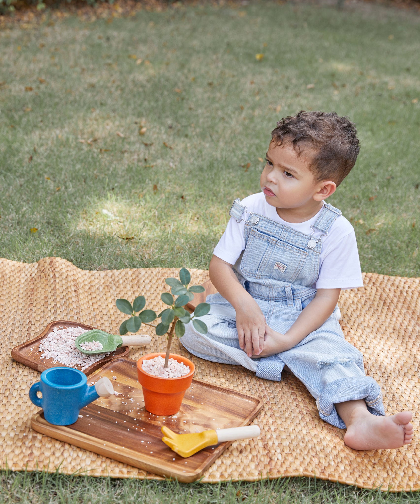 A child sitting on a mat outdoors playing with the A child playing with the PlanToys Gardening Set.