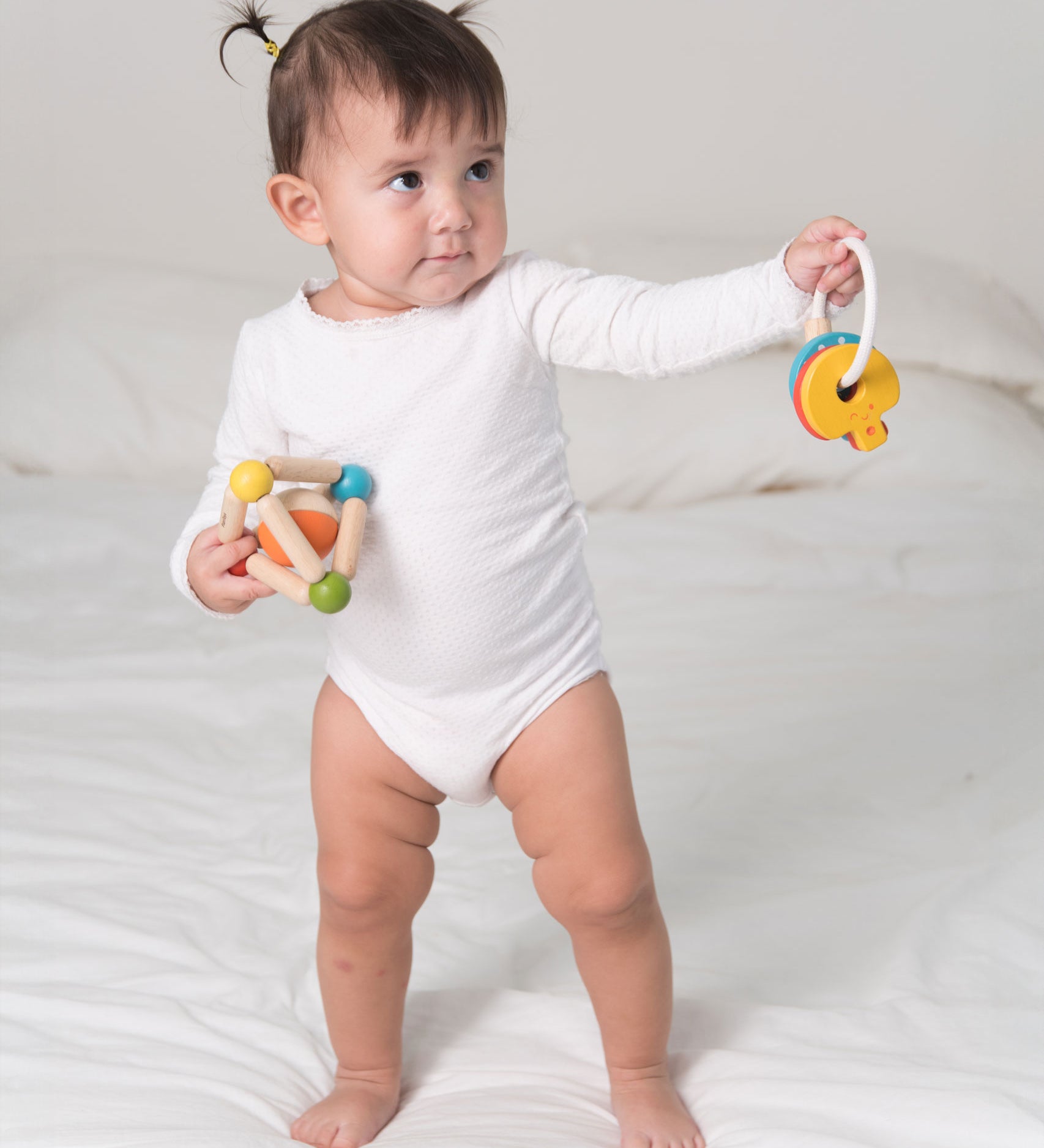 A toddler standing on a bed holding a PlanToys Triangle Clutching Toy in one hand and the key rattle in the other. 