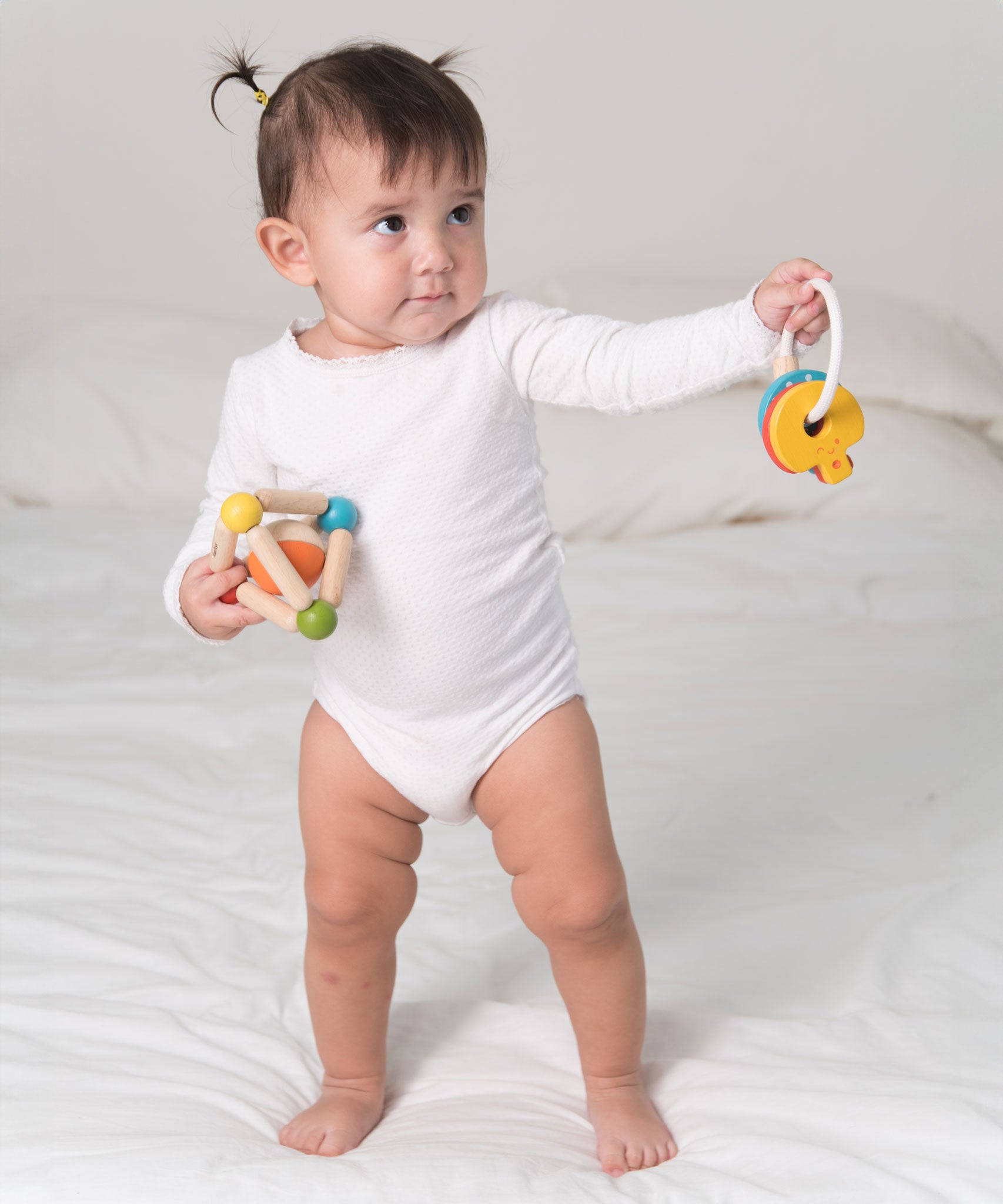 A toddler standing on a bed holding a PlanToys Triangle Clutching Toy in one hand and the key rattle in the other. 