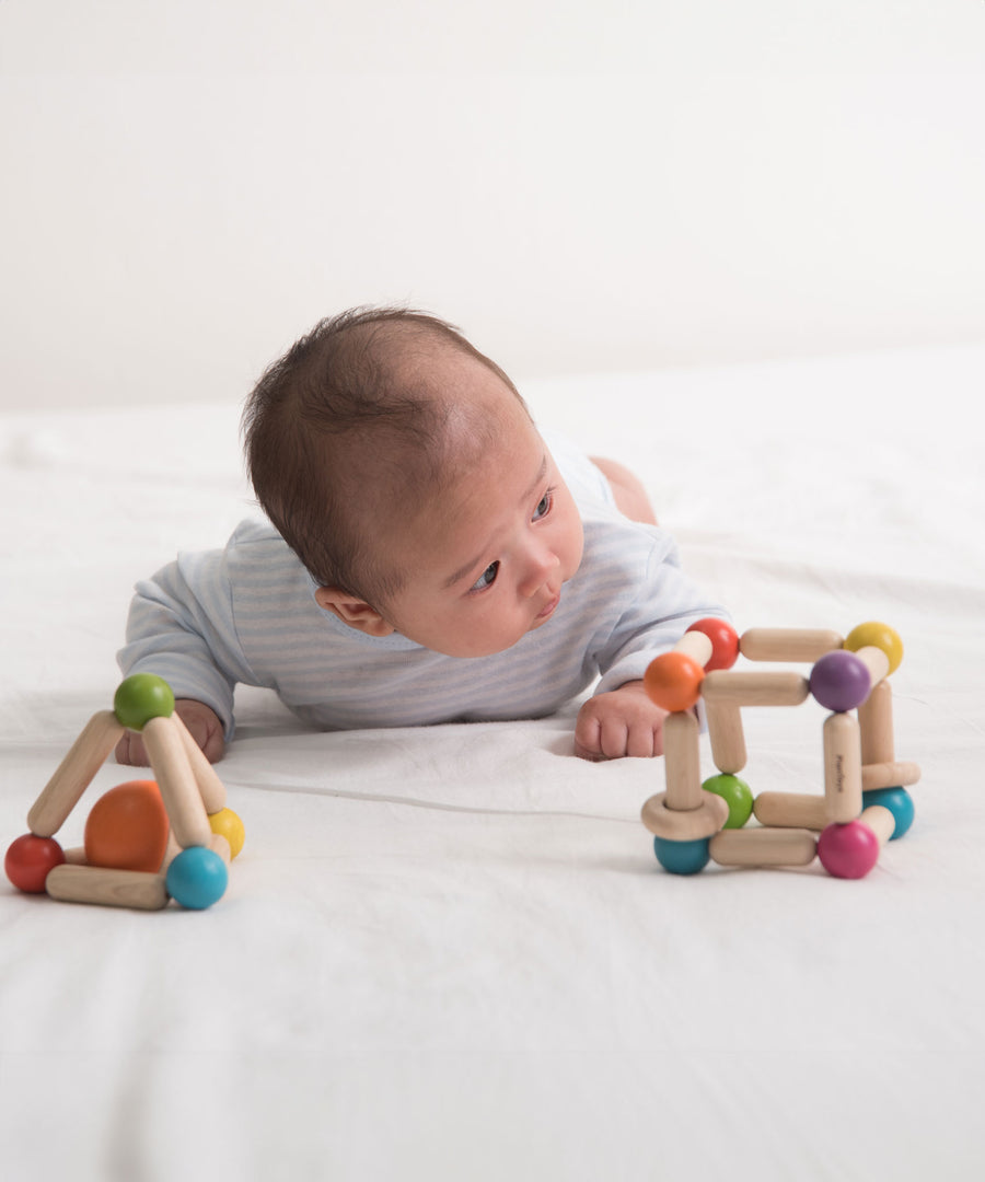 A baby is laying on his front on a bed with PlanToys Triangle and cube Clutching Toys in front of them. 