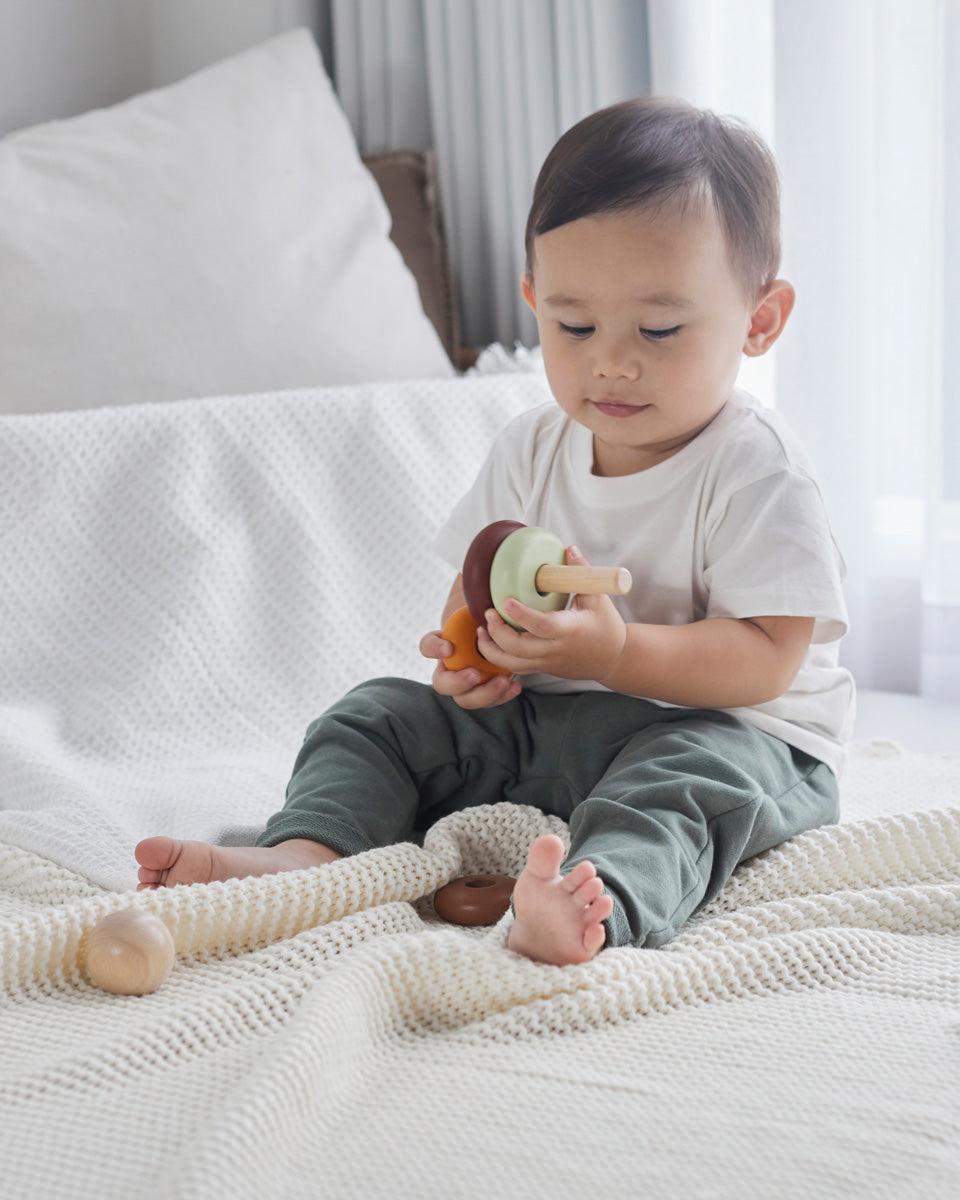 Young boy sat on a white bed playing with the PlanToys wooden stacking rings Waldorf toy