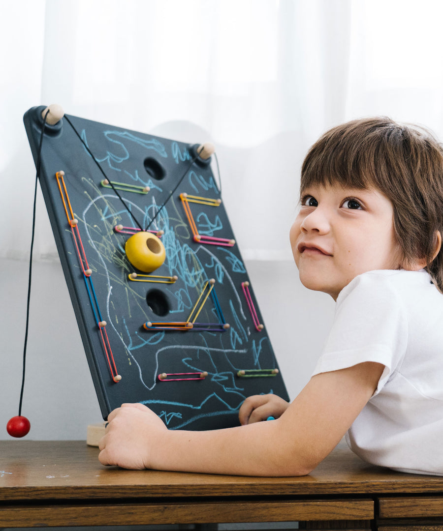 A child laying in front of a the PlanToys Wall Ball Game. The child has decorated the board with drawings. 
