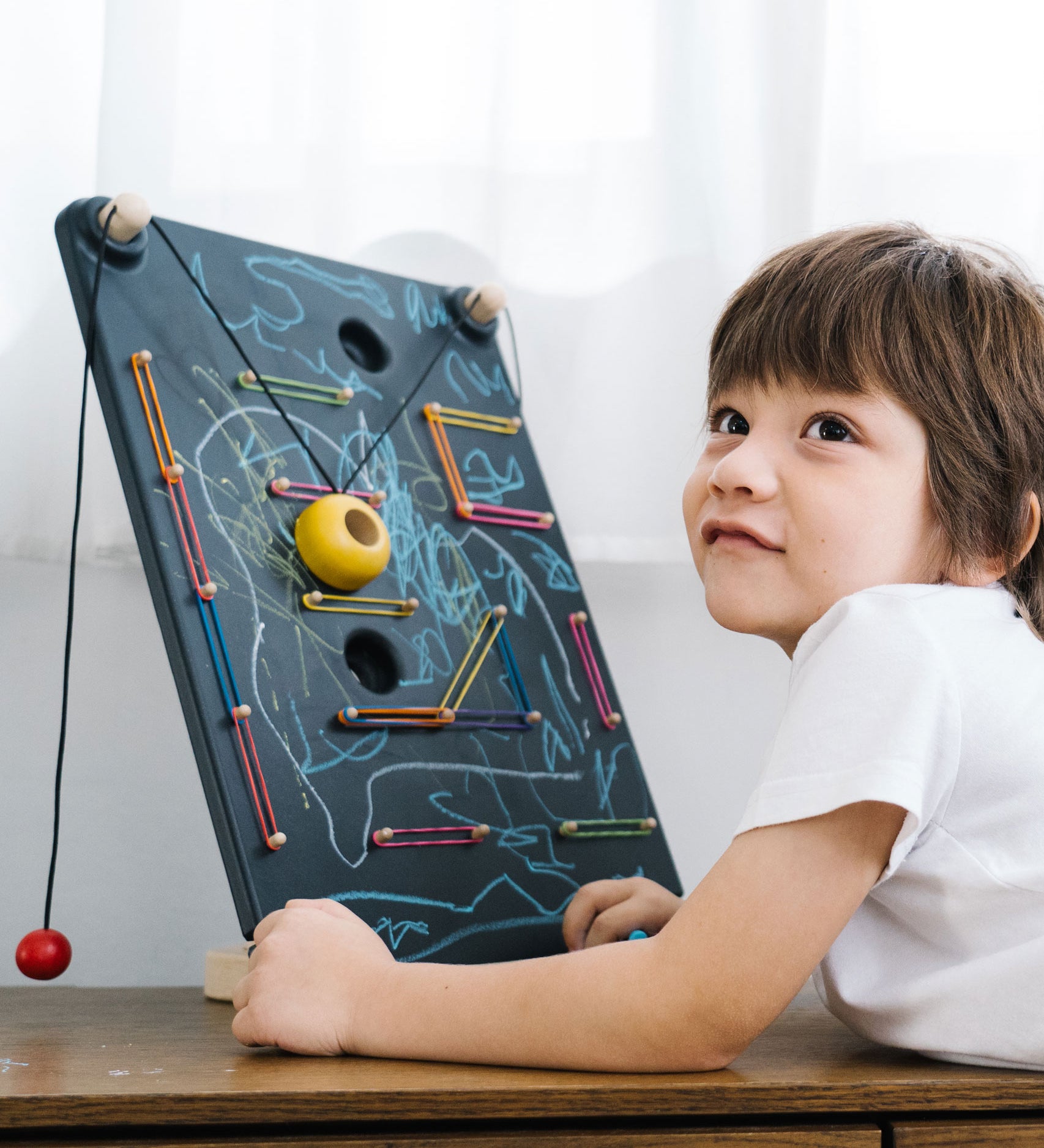 A child laying in front of a the PlanToys Wall Ball Game. The child has decorated the board with drawings. 
