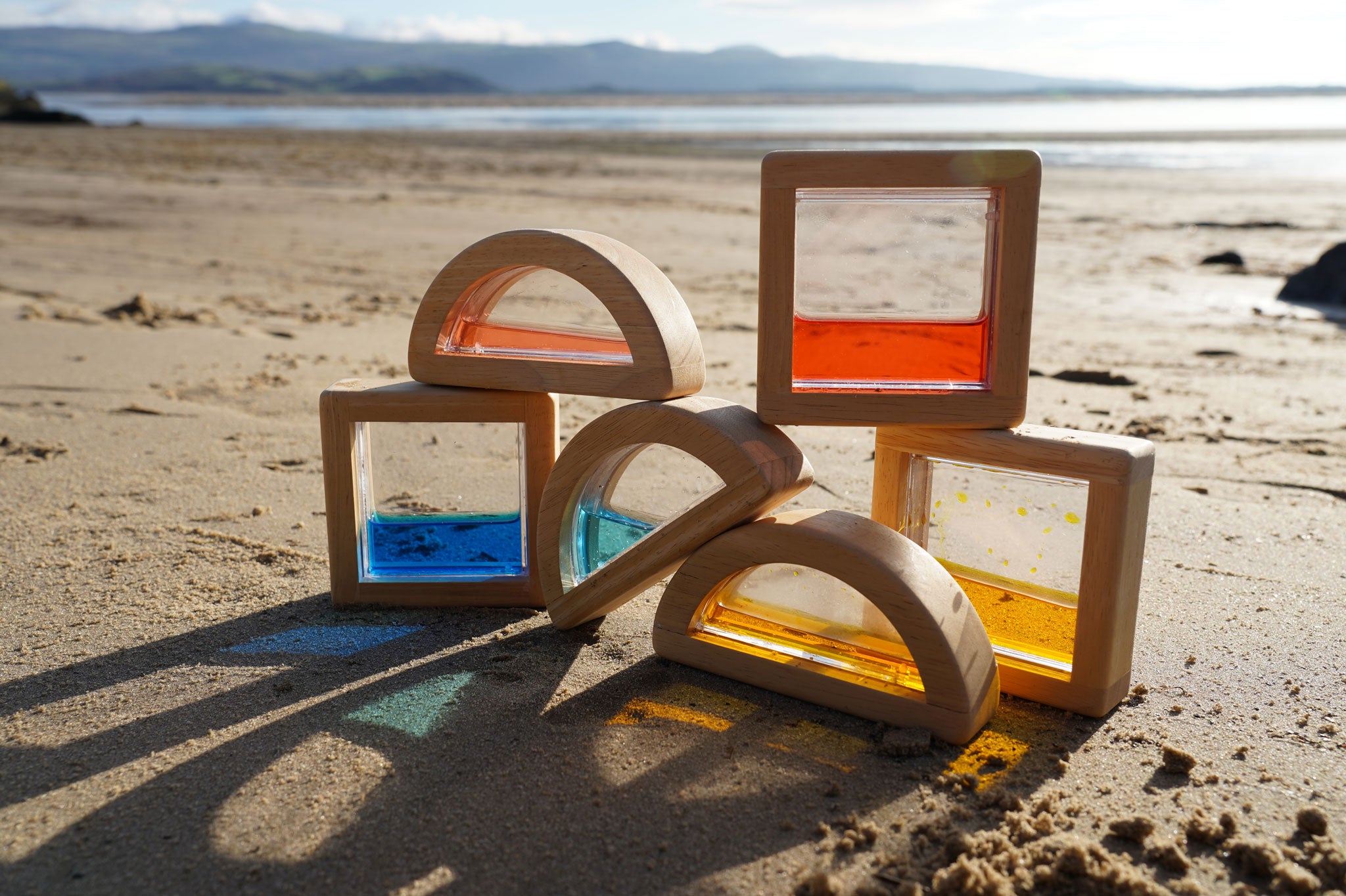 The PlanToys Water Blocks placed on sand on a beach. The light is shining through the blocks casting shadows and colours on the sand. 
