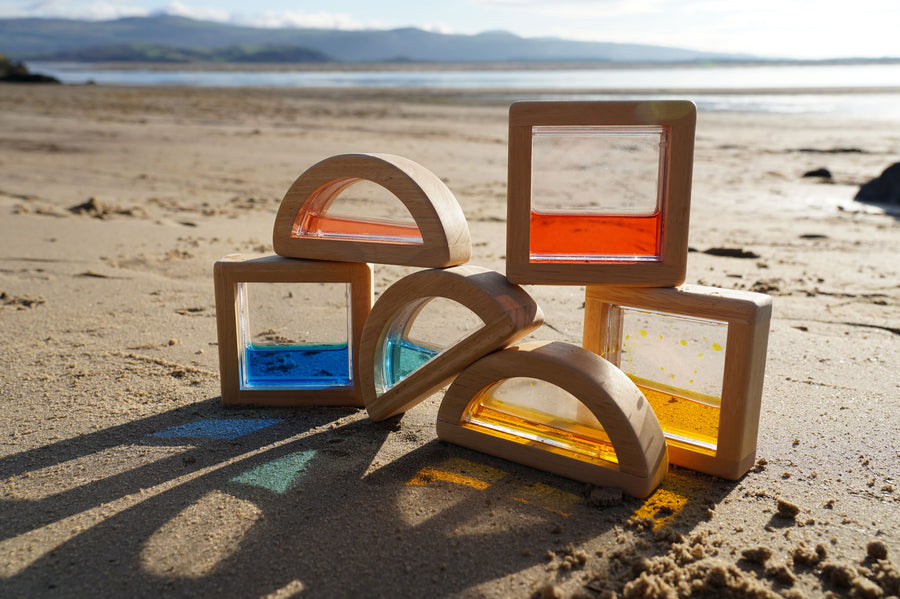 The PlanToys Water Blocks placed on sand on a beach. The light is shining through the blocks casting shadows and colours on the sand. 
