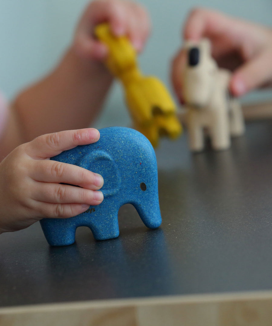 A child playing with the PlanToys Wild Animals Set, a child's hand can be seen holding the blue coloured elephant figure with the other animal figures in the background,