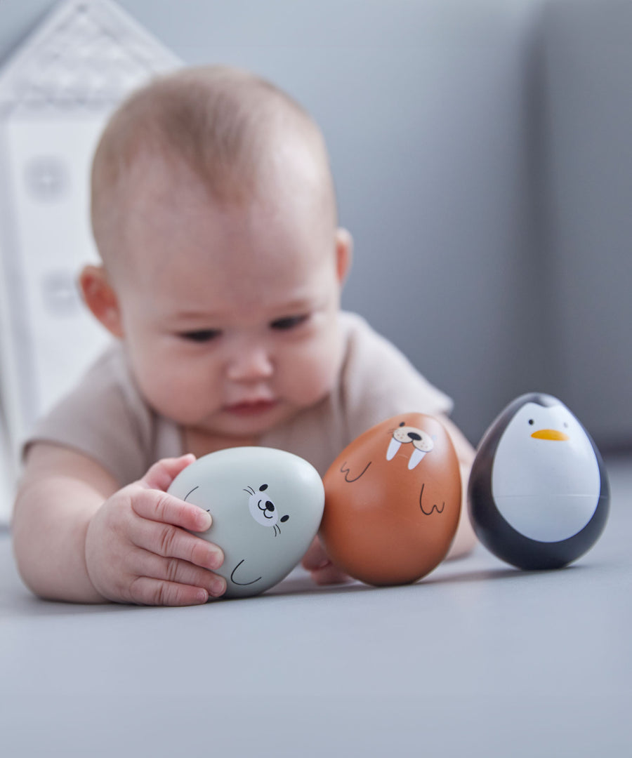 A baby laying on a grey floor reaching for the PlanToys wobbly seal, the walrus and penguin wobbly toys are besides them. 