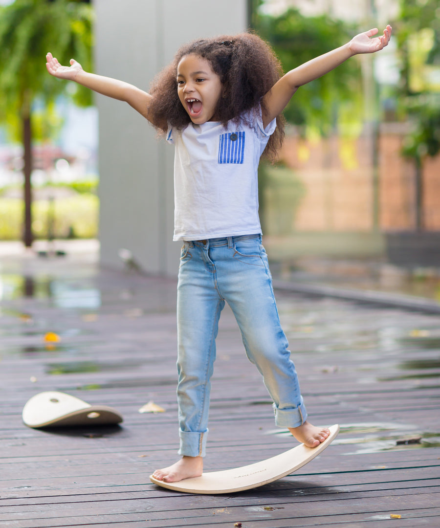 A child standing on the PlanToys Balance Board with both their arms help up high in a y shape. The board has been positioned on wooden decking outdoors. 