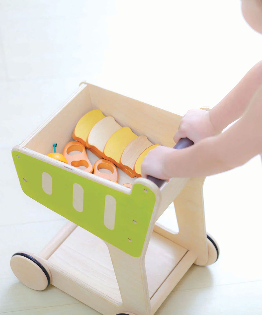 A PlanToys Shopping Trolley on a white surface, a child's hand can be seen pushing the trolley along. 