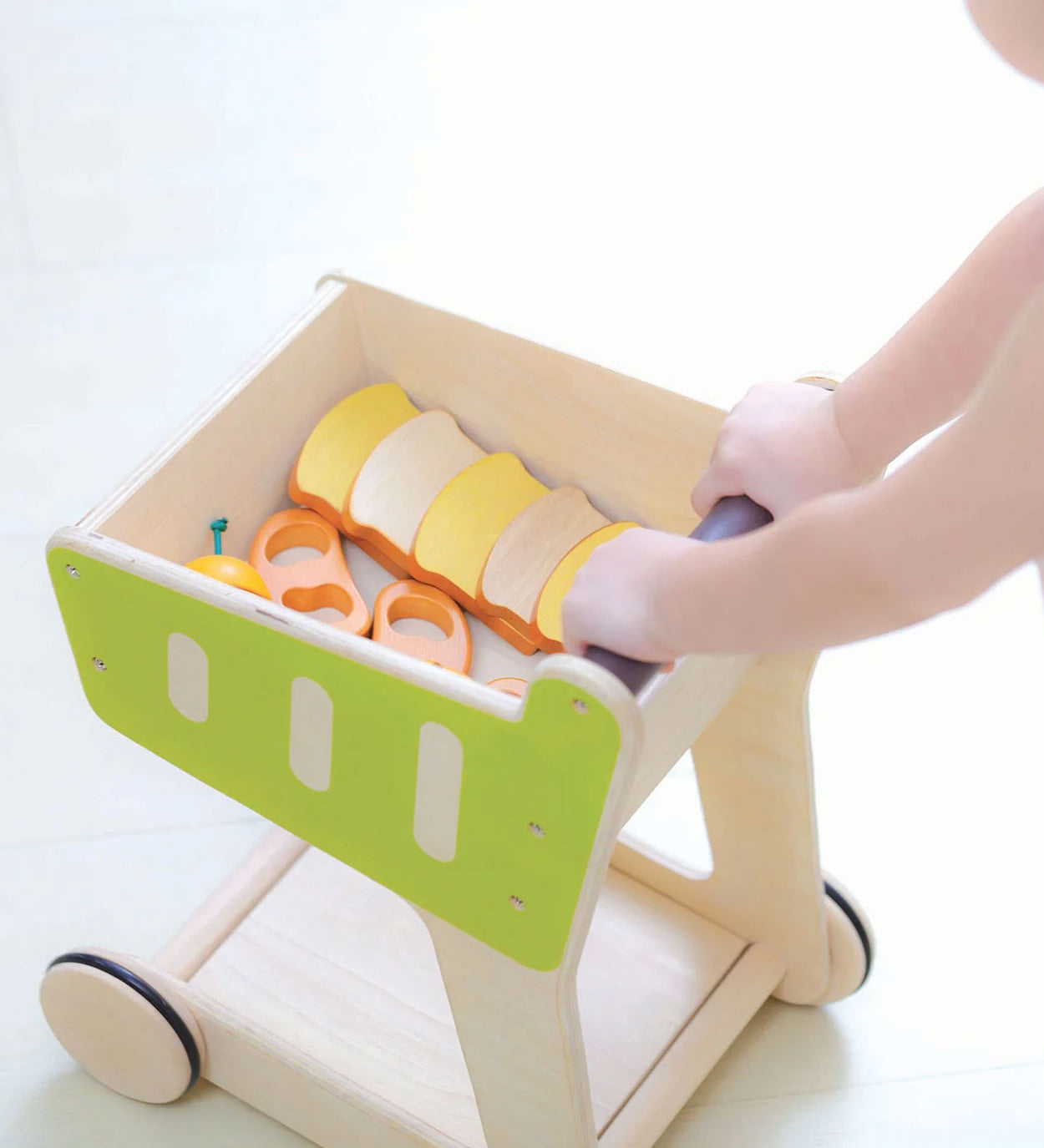 A PlanToys Shopping Trolley on a white surface, a child's hand can be seen pushing the trolley along. 