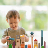 A child playing with the PlanToys Wooden Cityscape Blocks, the child is holding one of the blue blocks in one hand and a yellow one in the other. 