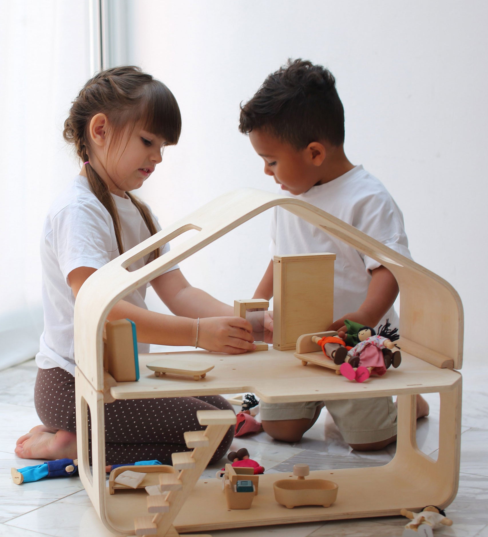 Two children playing with the PlanToys Contemporary Dolls House. There are wooden dolls and wooden furniture inside. 