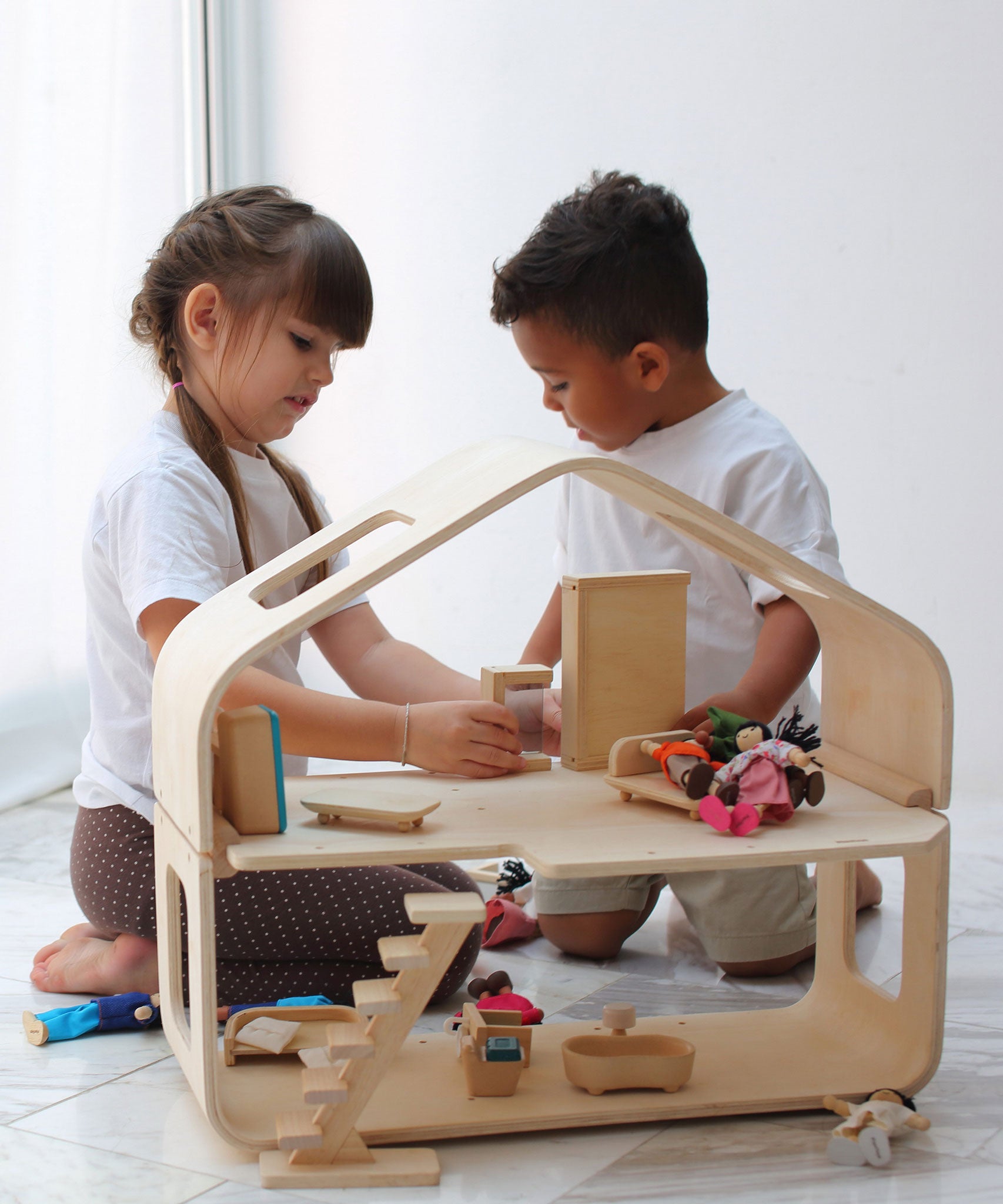 Two children playing with the PlanToys Contemporary Dolls House. There are wooden dolls and wooden furniture inside. 