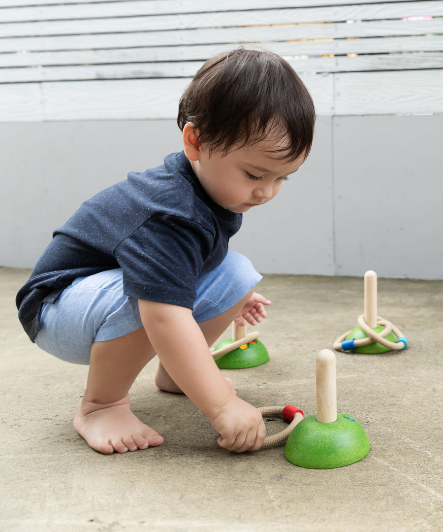 A child playing with the PlanToys Meadow Ring Toss they are kneeling down picking up one of the rings off the ground. 
