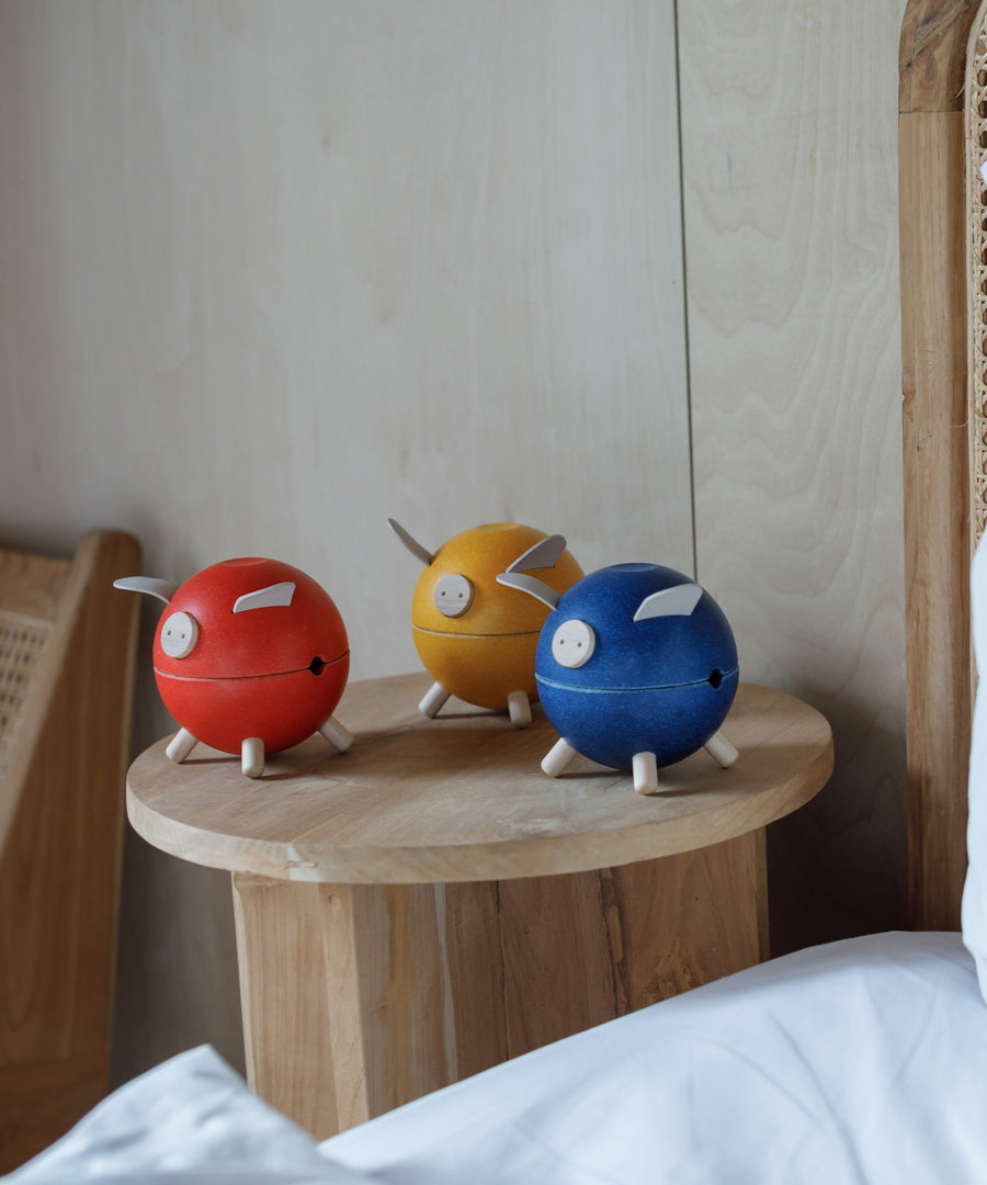 Three PlanToys piggy banks on a wooden side table. One red, one yellow and one blue. 