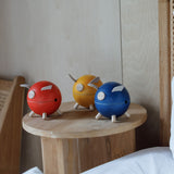 Three PlanToys piggy banks on a wooden side table. One red, one yellow and one blue. 