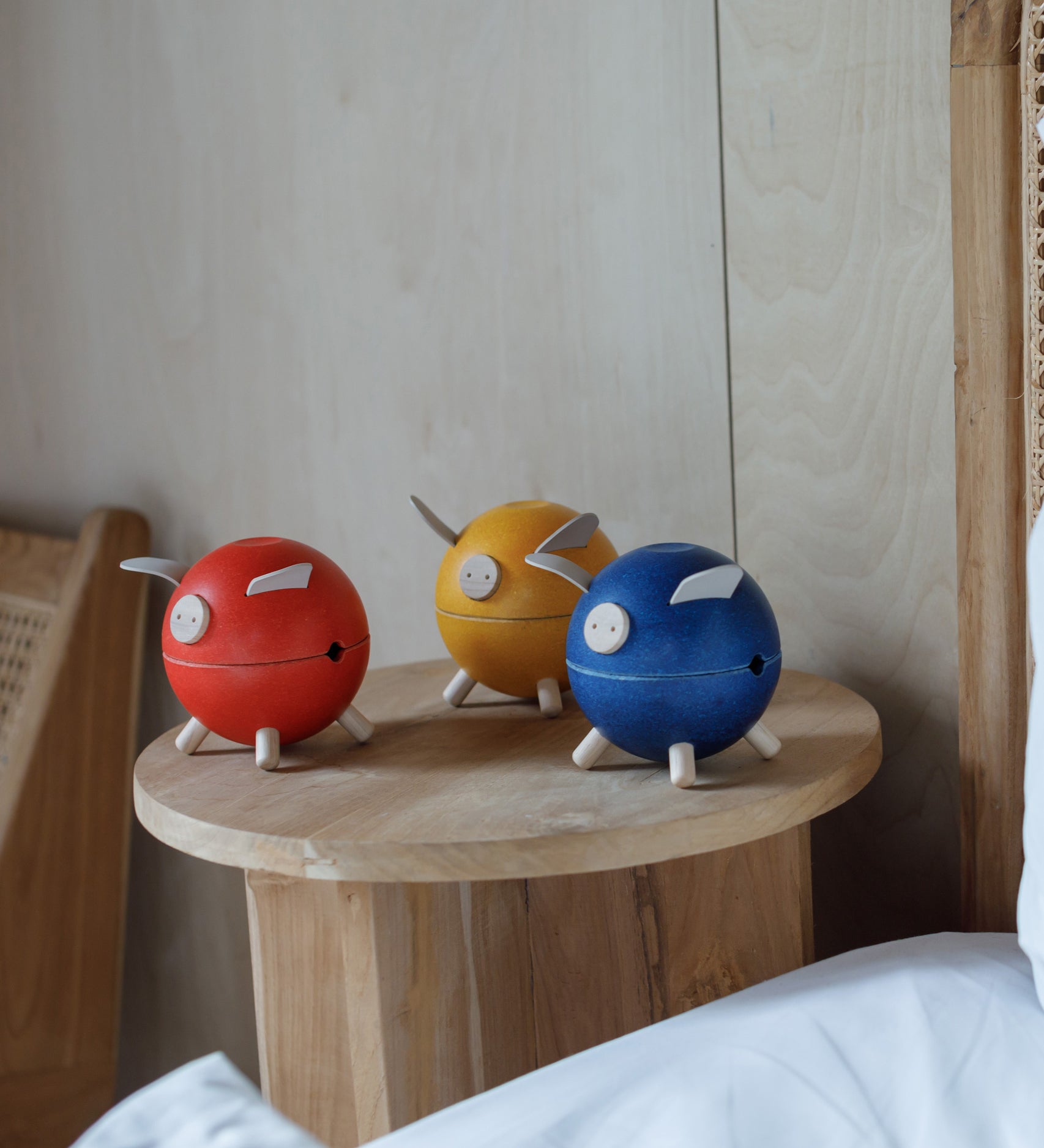 Three PlanToys piggy banks on a wooden side table. One red, one yellow and one blue. 