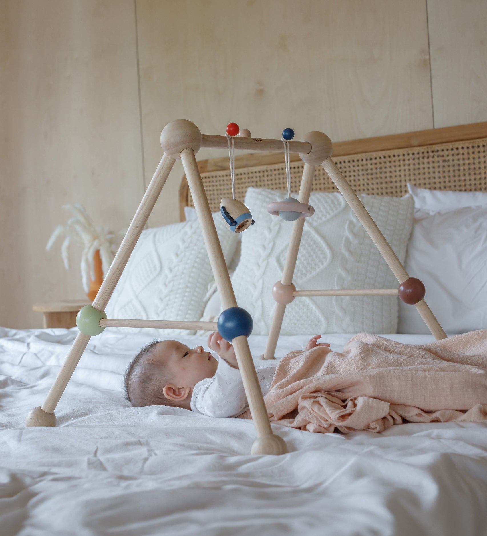 A baby laying underneath the PlanToys Play Gym Orchard on a bed which is covered in crisp white linen. 