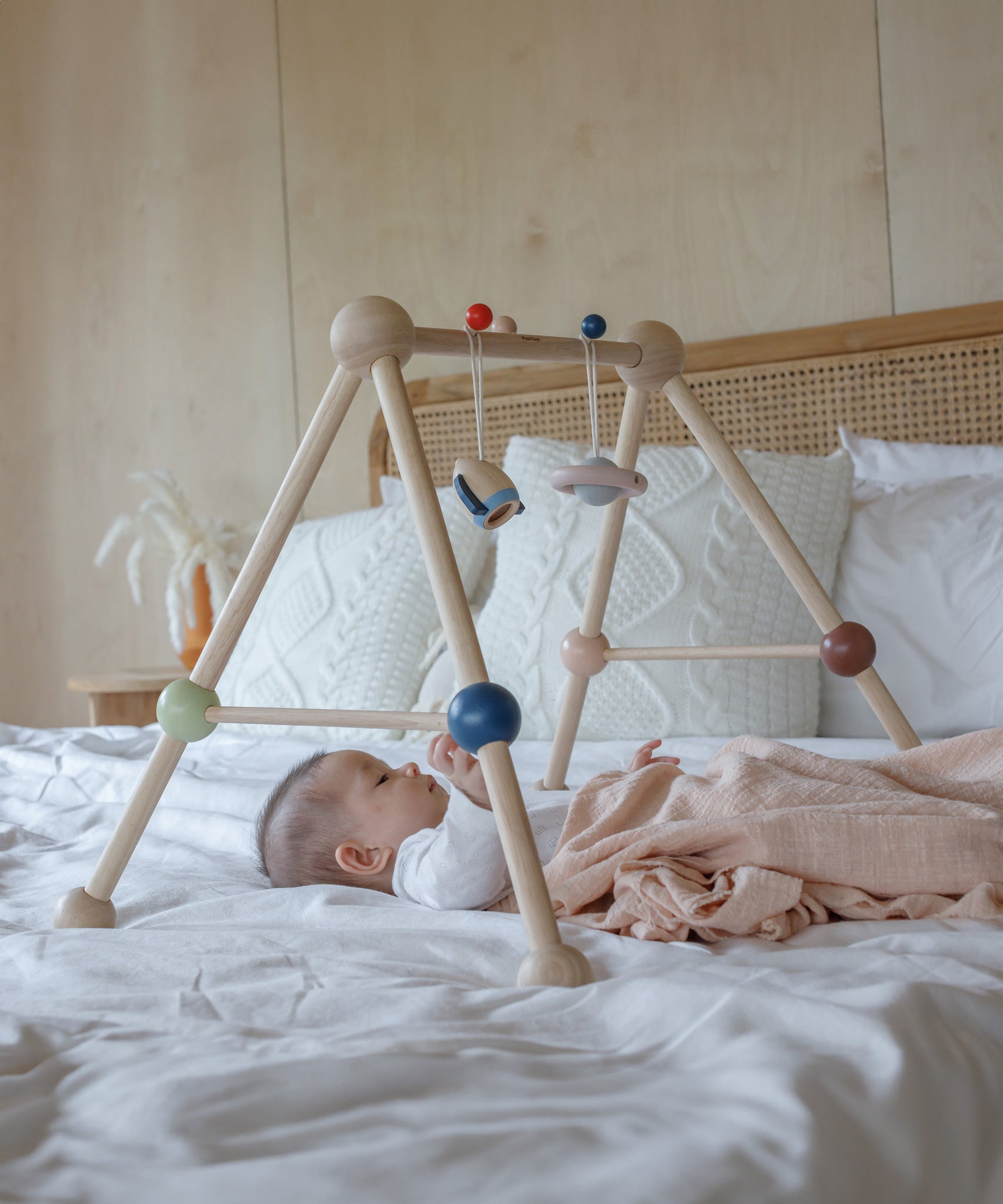 A baby laying underneath the PlanToys Play Gym Orchard on a bed which is covered in crisp white linen. 