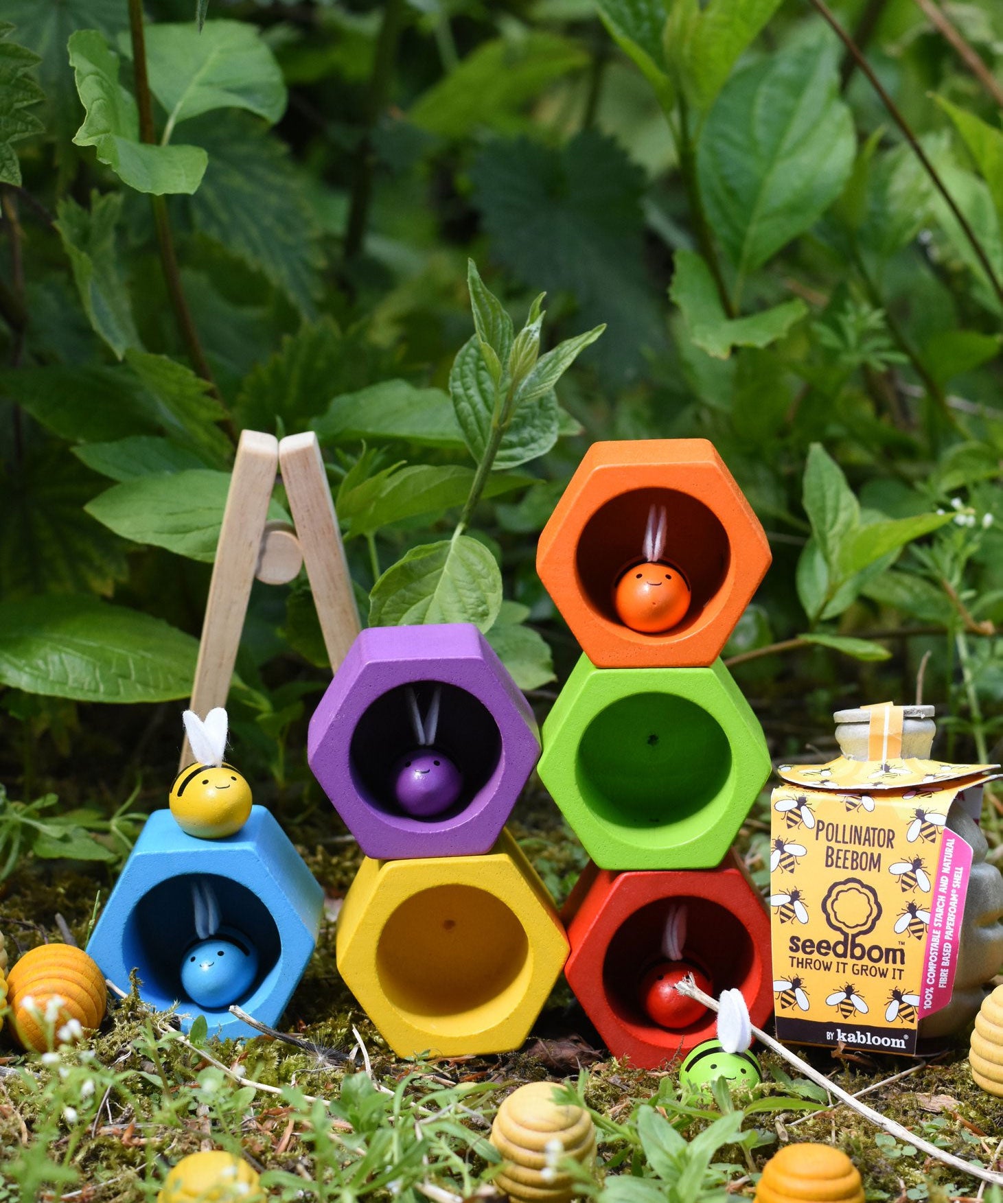 The Plan Toys Bee Hives Rainbow hives stacked up outdoors, some of the bees are in the matching coloured hives. There are Grapat honeycomb pieces placed in front of them and a pollinator seedbom by the side. 