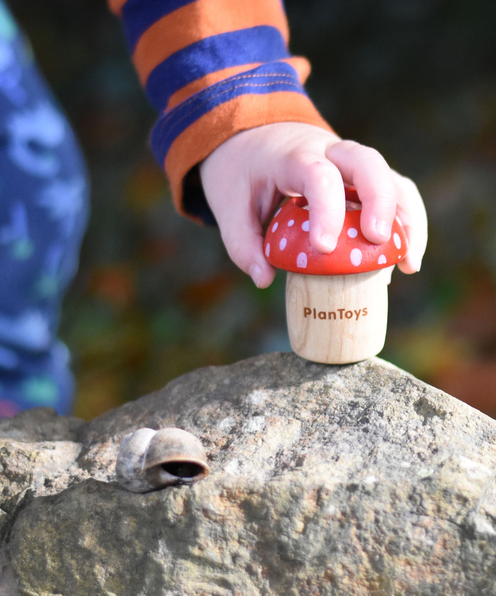 A child's hand reaching for a red PlanToys Mushroom Kaleidoscope that has been placed on a large rock. 