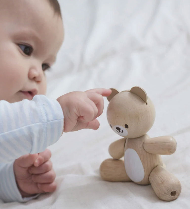 A baby playing with a PlanToys wooden toy bear on a white material. 