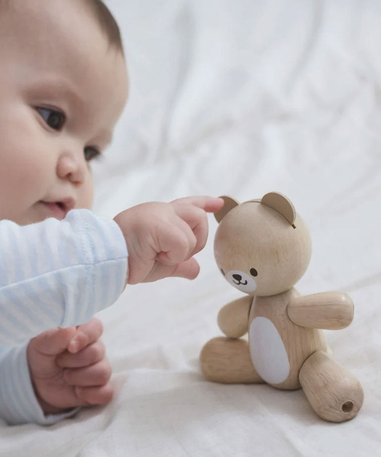 A baby playing with a PlanToys wooden toy bear on a white material. 